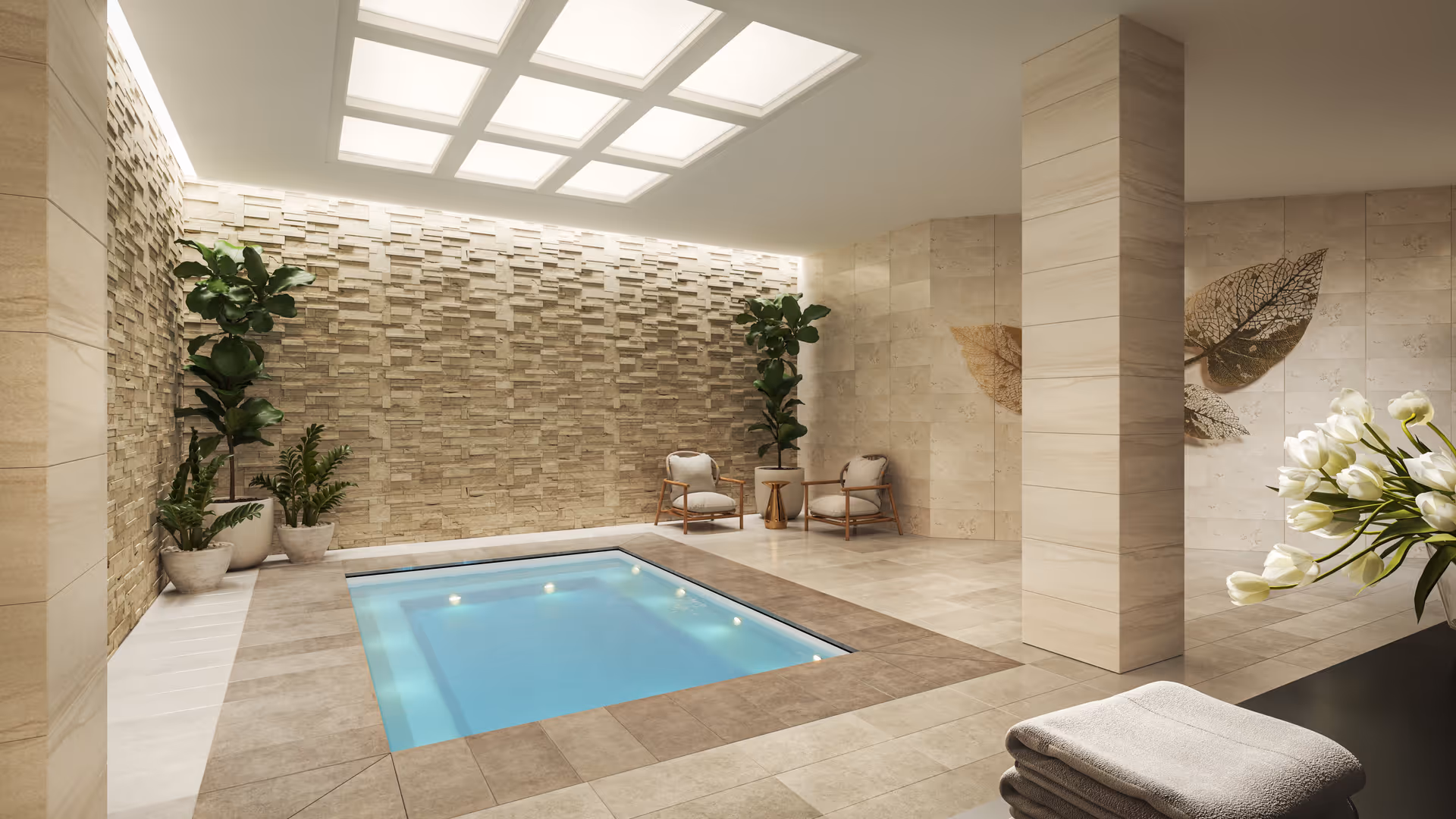 Indoor spa area with a small rectangular pool surrounded by beige tiled flooring and walls. There are two cushioned chairs with a small table between them, several potted plants, decorative leaf wall art, and a stack of folded towels on a black surface. The ceiling features recessed lighting panels.