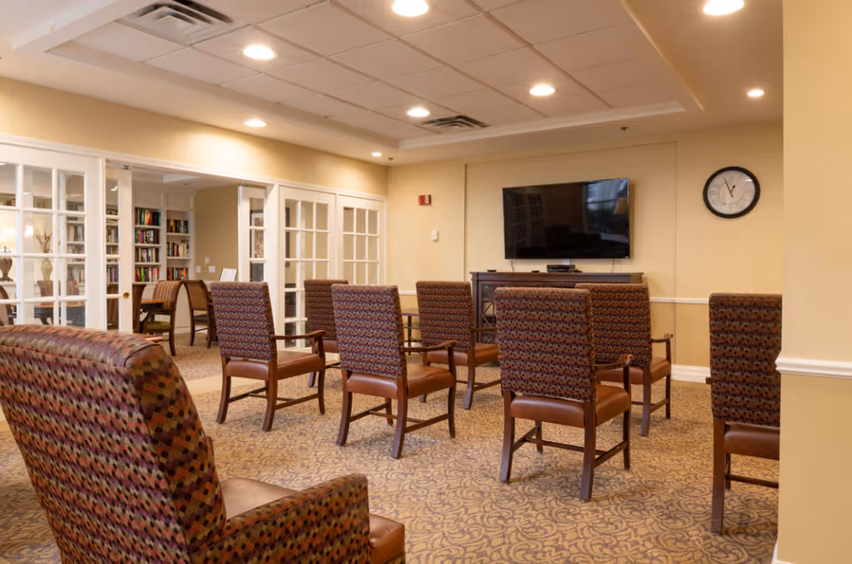 A cozy common area with multiple patterned armchairs arranged in rows facing a wall-mounted flat-screen TV. The room has beige walls, a patterned carpet, recessed ceiling lights, and a clock on the wall. There are glass-paneled double doors leading to another room with bookshelves and additional seating.