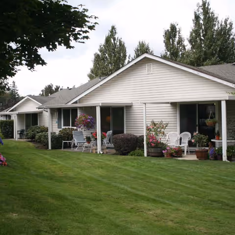 Single-story residential building with beige siding and a gray roof, featuring small covered patios with outdoor chairs, potted plants, and hanging flower baskets. The building is surrounded by a well-maintained green lawn and trees in the background under a cloudy sky.