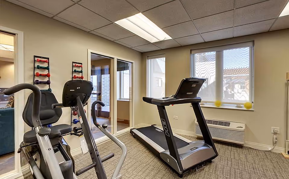 A small fitness room with exercise equipment including a treadmill and a stationary bike. There are two windows with blinds partially drawn, a wall-mounted rack holding colorful dumbbells, and a heating/cooling unit below the windows. The room has beige walls and a carpeted floor.
