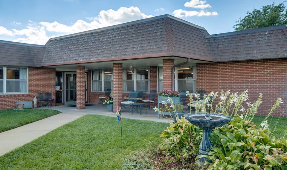 Exterior view of a brick building with a covered patio area furnished with chairs and tables. There is a well-maintained lawn and garden with a birdbath in the foreground under a partly cloudy sky.