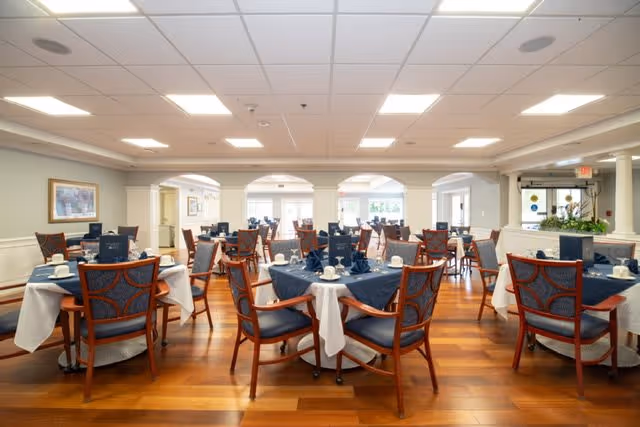 A spacious dining room with multiple round tables covered with white and navy blue tablecloths, each set with cups, saucers, and napkins. The room features wooden chairs with blue cushioned seats and backs, hardwood floors, and large windows allowing natural light to fill the space. The walls are light-colored with white trim, and there are decorative arches and framed artwork on the walls.
