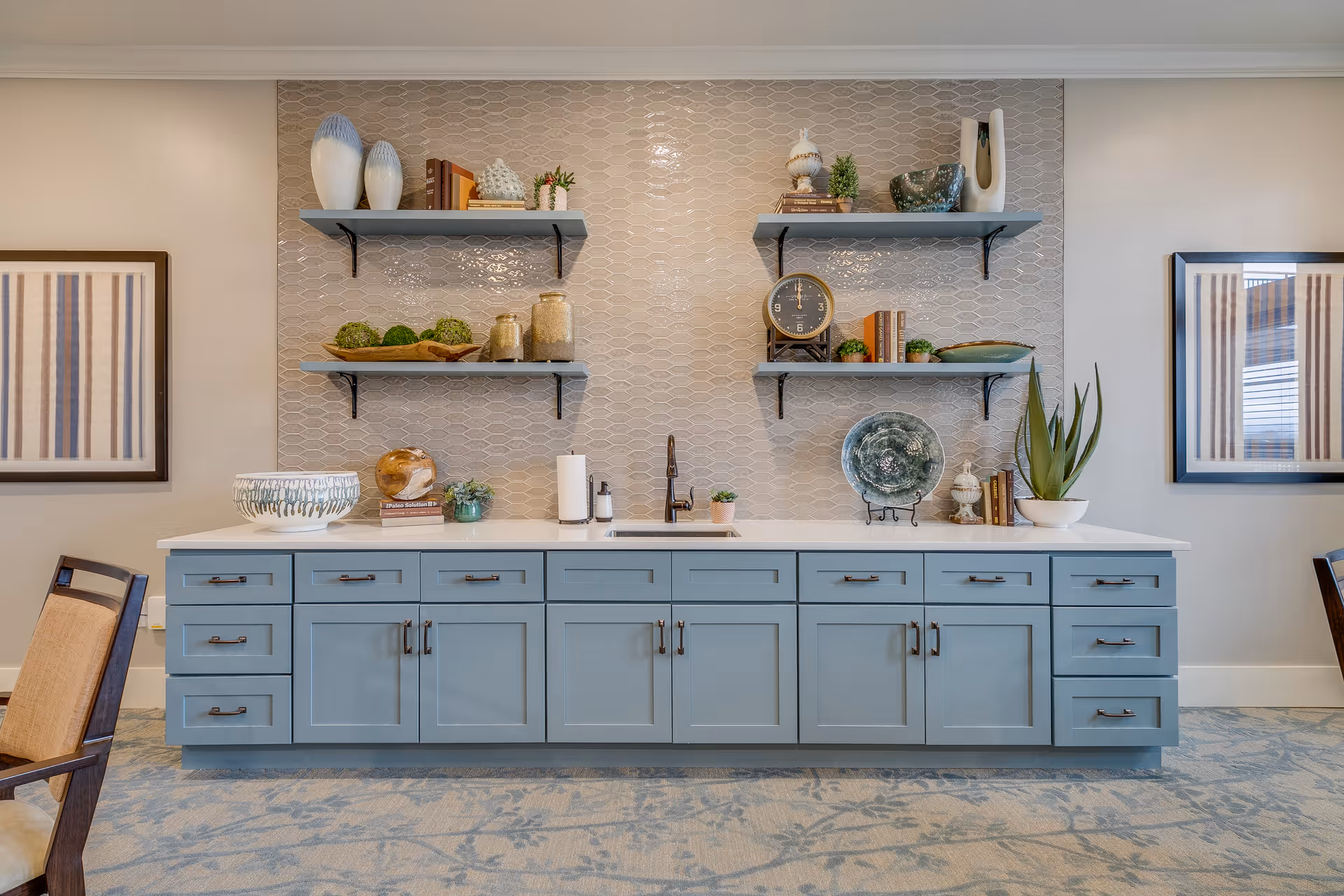 A modern kitchen area with light blue cabinets and a white countertop. Above the countertop are two sets of floating shelves decorated with vases, books, plants, jars, a clock, and decorative plates. There is a small sink with a black faucet in the center of the countertop. On either side of the kitchen area are framed striped artworks on the wall. The floor has a patterned carpet in shades of blue and beige.