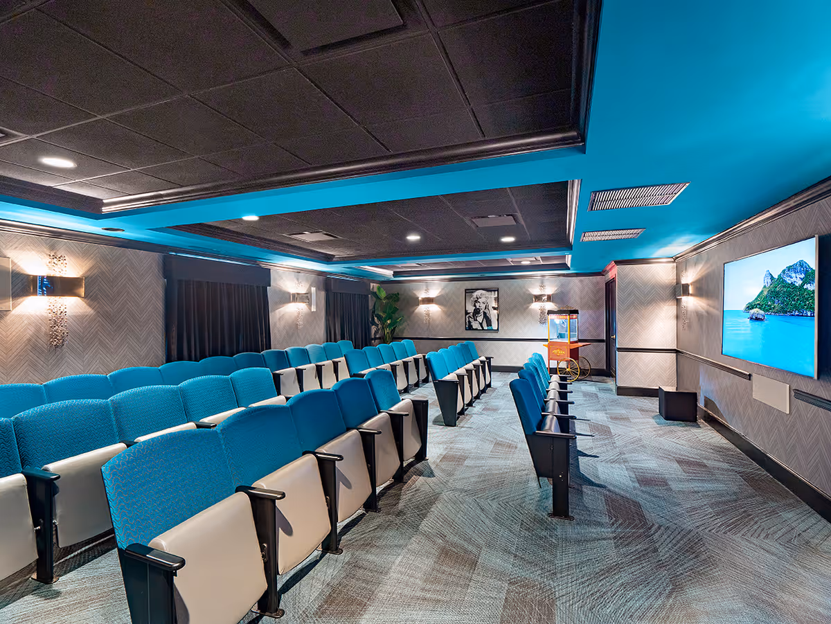 A theater-style room with rows of blue and beige cushioned seats facing a large flat-screen TV mounted on the wall. The room has a patterned carpet, gray walls with wall sconces, and a ceiling with blue accent lighting. There is a popcorn machine in the back corner and a black and white portrait on the wall.