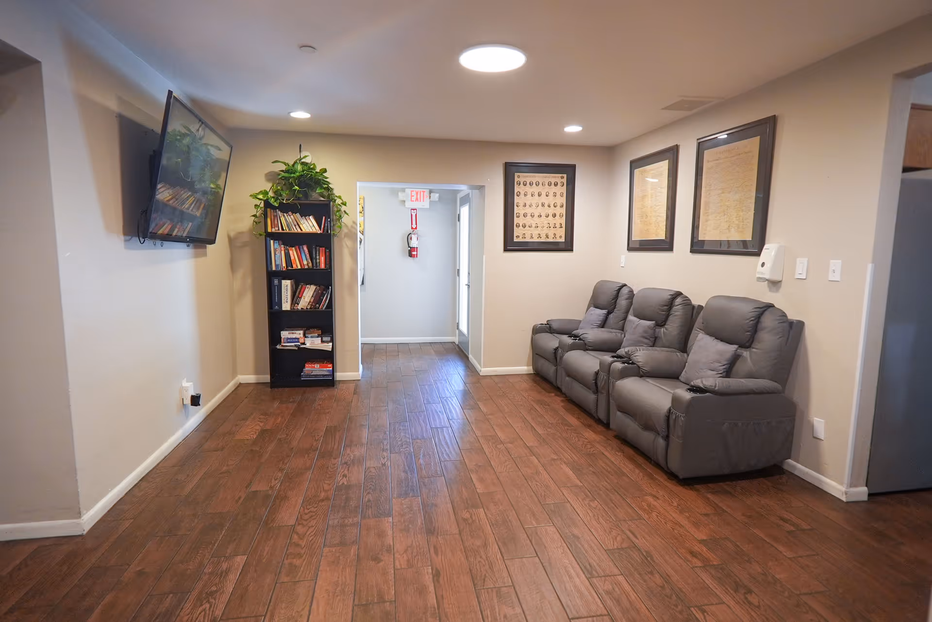 A small lounge area with three gray recliner chairs against a beige wall adorned with framed pictures. A black bookshelf filled with books and a green plant on top is positioned near a doorway with an exit sign and fire extinguisher visible. A flat-screen TV is mounted on the opposite wall. The floor is covered with wood-like tiles and the ceiling has recessed lighting.