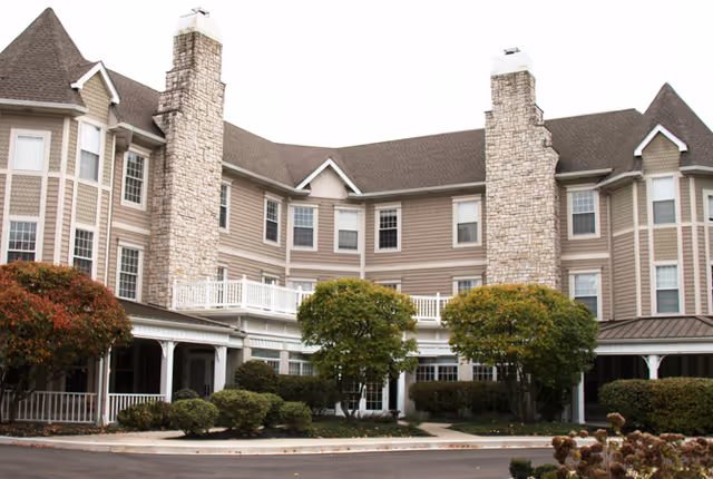 Exterior view of a multi-story senior living facility with beige siding and stone chimneys. The building features multiple windows, a covered porch area, and landscaped bushes and trees in front.