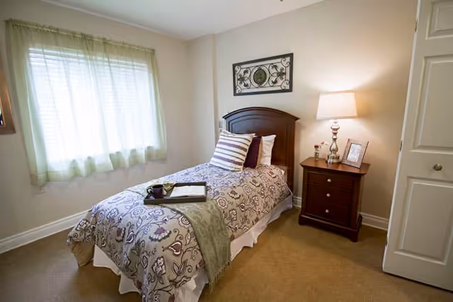 A cozy bedroom with a single bed covered in a patterned bedspread and pillows. A tray with a cup and an open book rests on the bed. Next to the bed is a wooden nightstand with a lamp, a small flower vase, and a framed photo. A window with sheer curtains lets in natural light, and a decorative wall piece hangs above the bed's headboard.