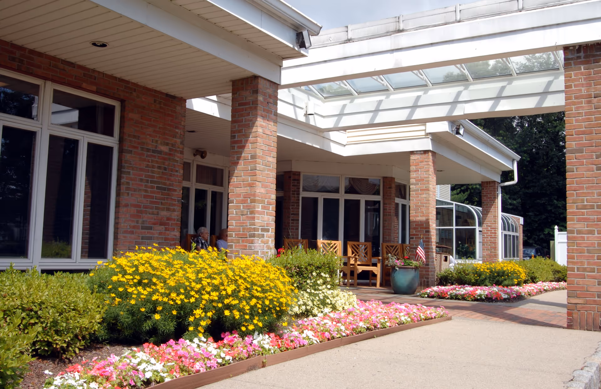 Outdoor patio area of a brick building with large windows, wooden chairs, and colorful flower beds featuring yellow, pink, and white flowers. Two people are seated on the patio under a covered walkway with a glass roof.