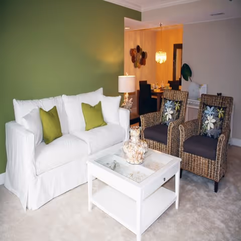 A cozy living room area with a white sofa adorned with two green pillows, two wicker armchairs with floral cushions, and a white coffee table with a decorative glass jar filled with seashells. In the background, a dining area with a table set for four and a hanging chandelier is visible.