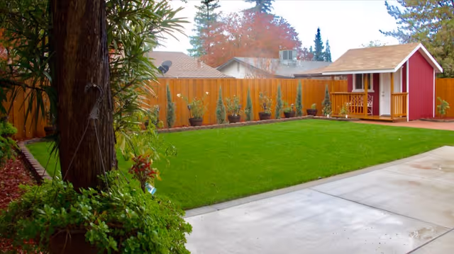 A backyard garden area with a well-maintained green lawn, a wooden fence lined with potted plants, a large tree with surrounding greenery on the left, and a small red and white playhouse or shed in the back right corner.