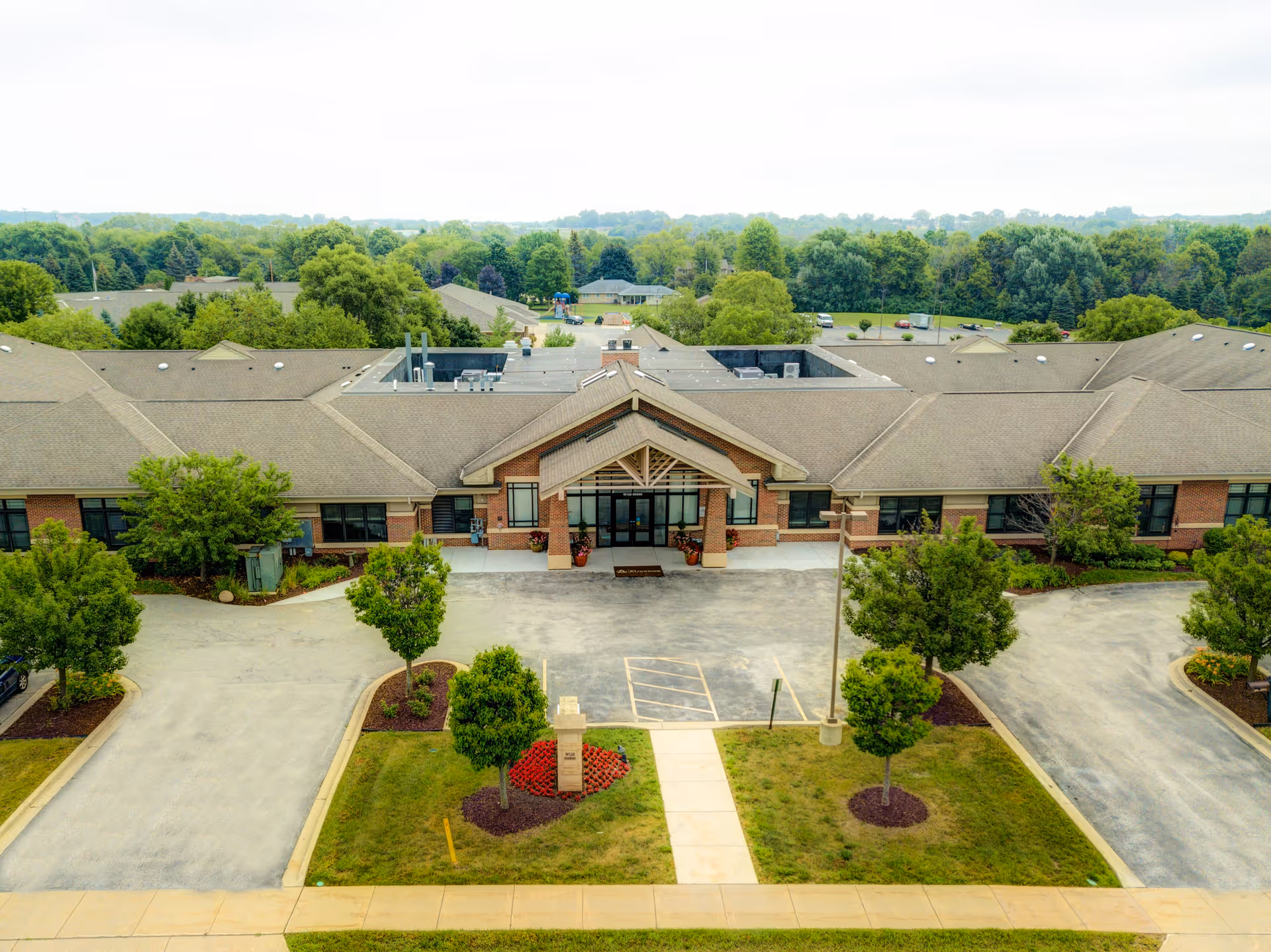 Aerial view of The Auberge at Oak Village single-story building with a covered main entrance, driveways, and landscaped grounds surrounded by trees.