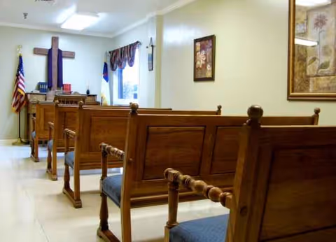 Interior room with wooden pews facing a front area with a cross and flags.