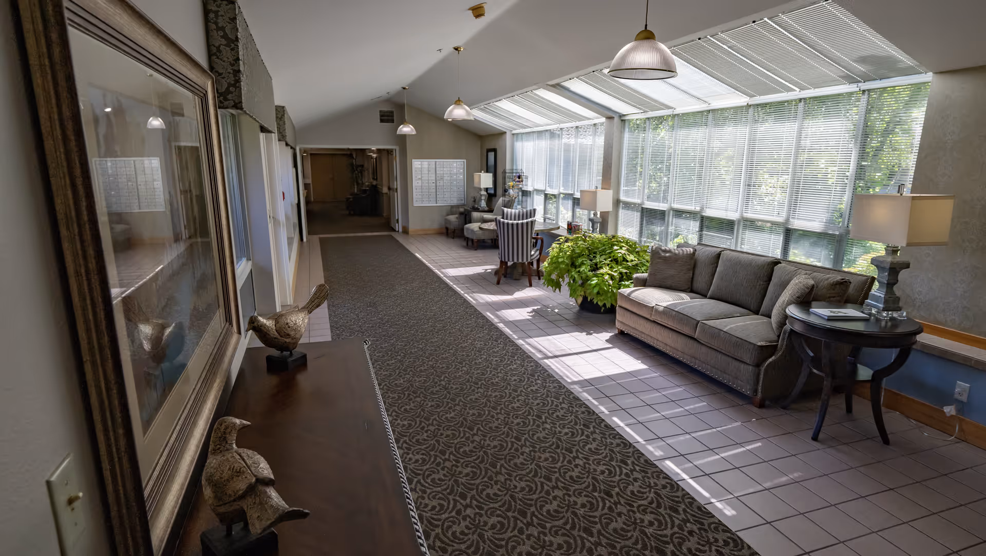 Sunlit hallway lounge with a sofa, chairs, side tables, plants and decorative bird sculptures on a console table.