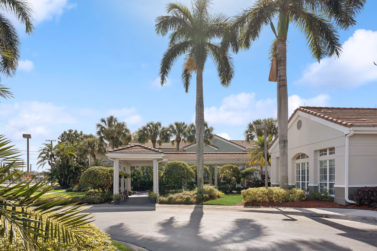 Entrance and porte-cochère of a senior living facility with palm trees and landscaped grounds under a blue sky.