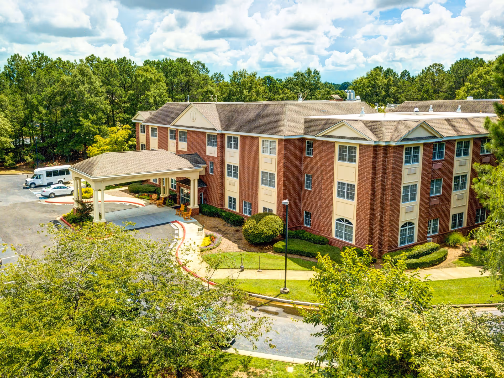 Exterior view of a three-story brick building with a covered entrance and surrounding greenery under a partly cloudy sky, identified as Magnolia Place of Roswell.