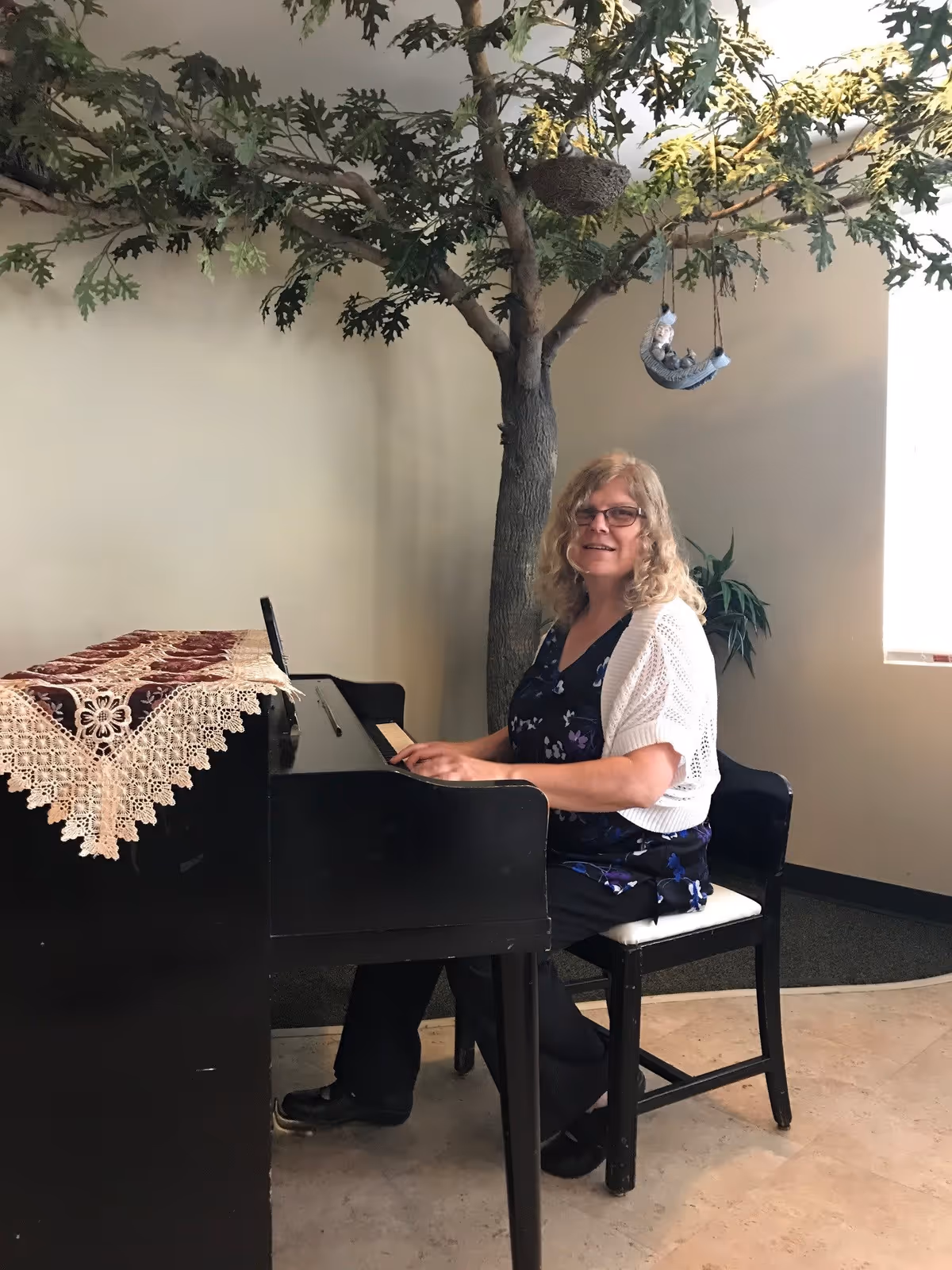 A woman with curly blonde hair and glasses sits on a chair playing a black piano in a room with a large artificial tree behind her. The piano has a decorative lace cloth on top, and there is a window letting in natural light on the right side.