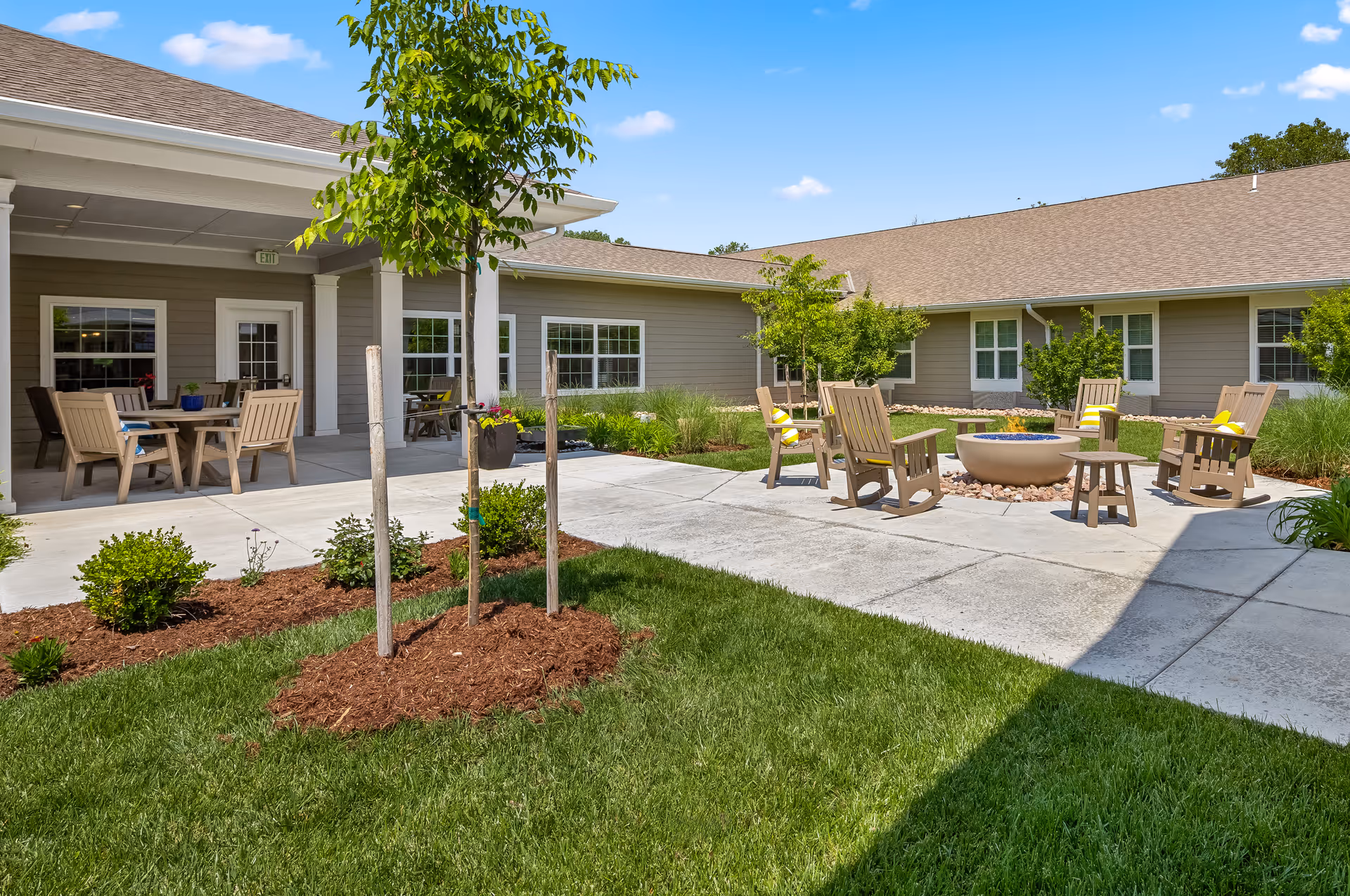 Outdoor courtyard area of a senior living facility with a fire pit surrounded by wooden chairs, small trees, green grass, and a covered patio with tables and chairs under a clear blue sky.