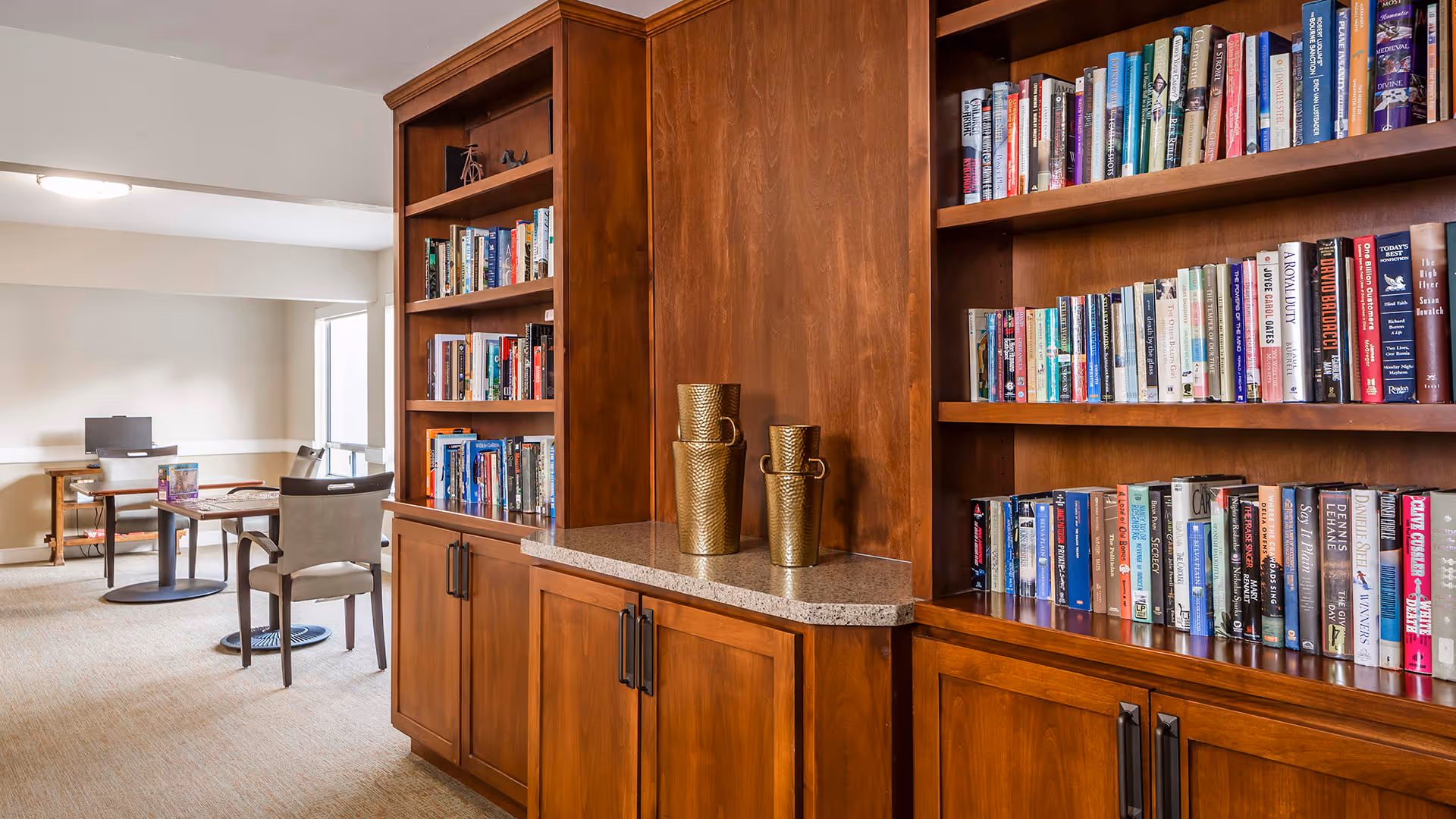 Interior view of a senior living facility library area with wooden bookshelves filled with books. There are two golden decorative vases on a granite countertop between the shelves. In the background, there are tables and chairs arranged for reading or activities in a well-lit room.