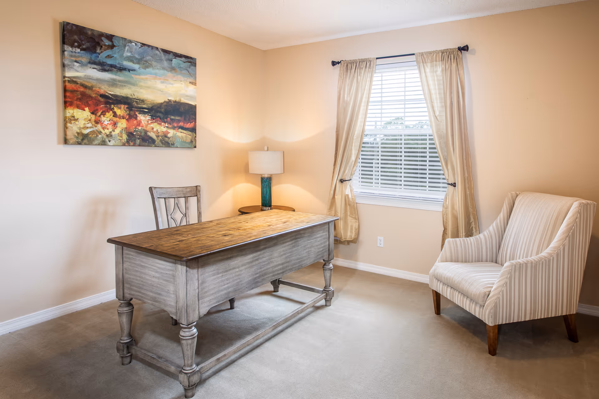 Bright furnished sitting room with a wooden desk, upholstered armchair, lamp, and a curtained window.