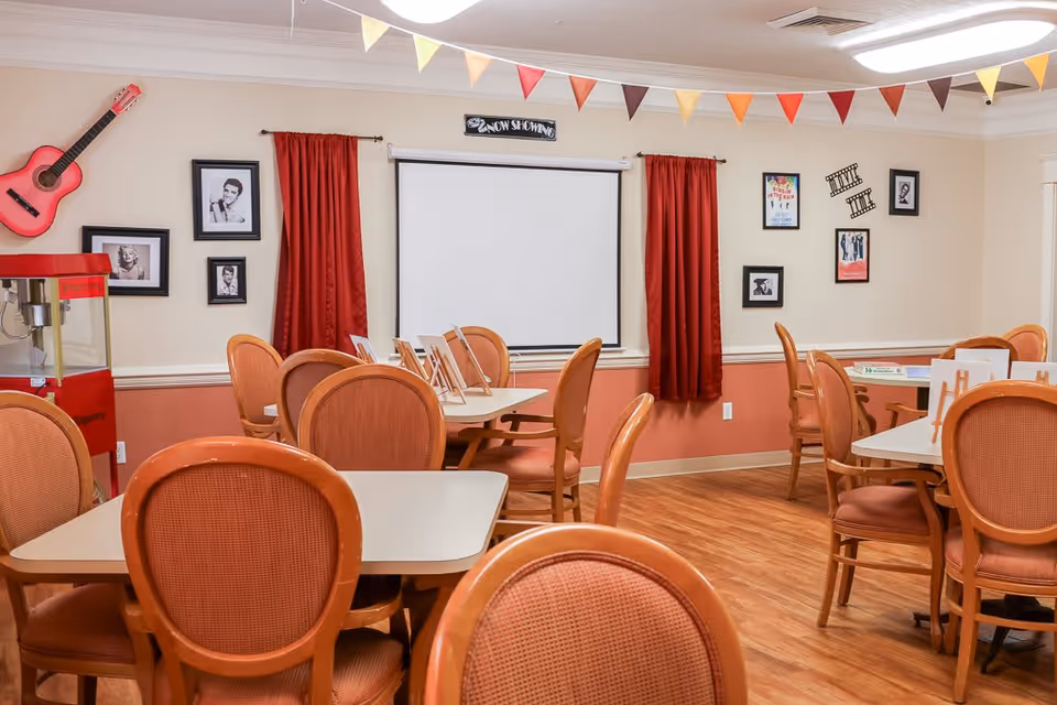 A cozy common room with several tables and orange cushioned chairs arranged around them. The walls are decorated with framed black-and-white photos, colorful posters, and a red guitar mounted on the wall. Red curtains flank a white projection screen with a 'Now Showing' sign above it. A red popcorn machine is visible on the left side, and colorful triangular pennant banners hang from the ceiling.