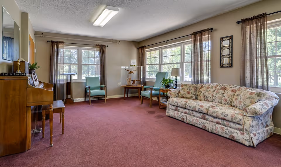 A cozy living room with a floral patterned sofa, two light blue armchairs, a wooden piano with a matching bench, and large windows with sheer brown curtains letting in natural light. The room has a red carpet and neutral-colored walls with minimal decor.