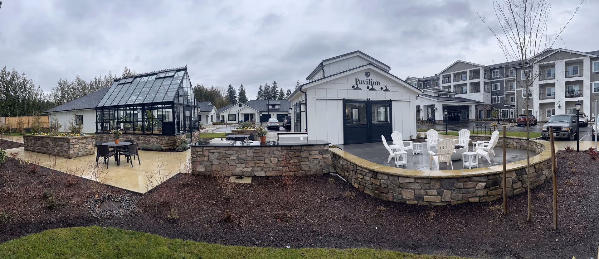 Outdoor area of The Inn at University Village Assisted Living Community featuring a glass greenhouse, a white pavilion building with black doors labeled 'Pavilion', stone walls, patio with white chairs around a fire pit, and a multi-story residential building in the background under a cloudy sky.