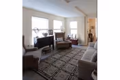 Bright living room with a sofa, two armchairs, patterned area rug, wooden sideboard and three windows.