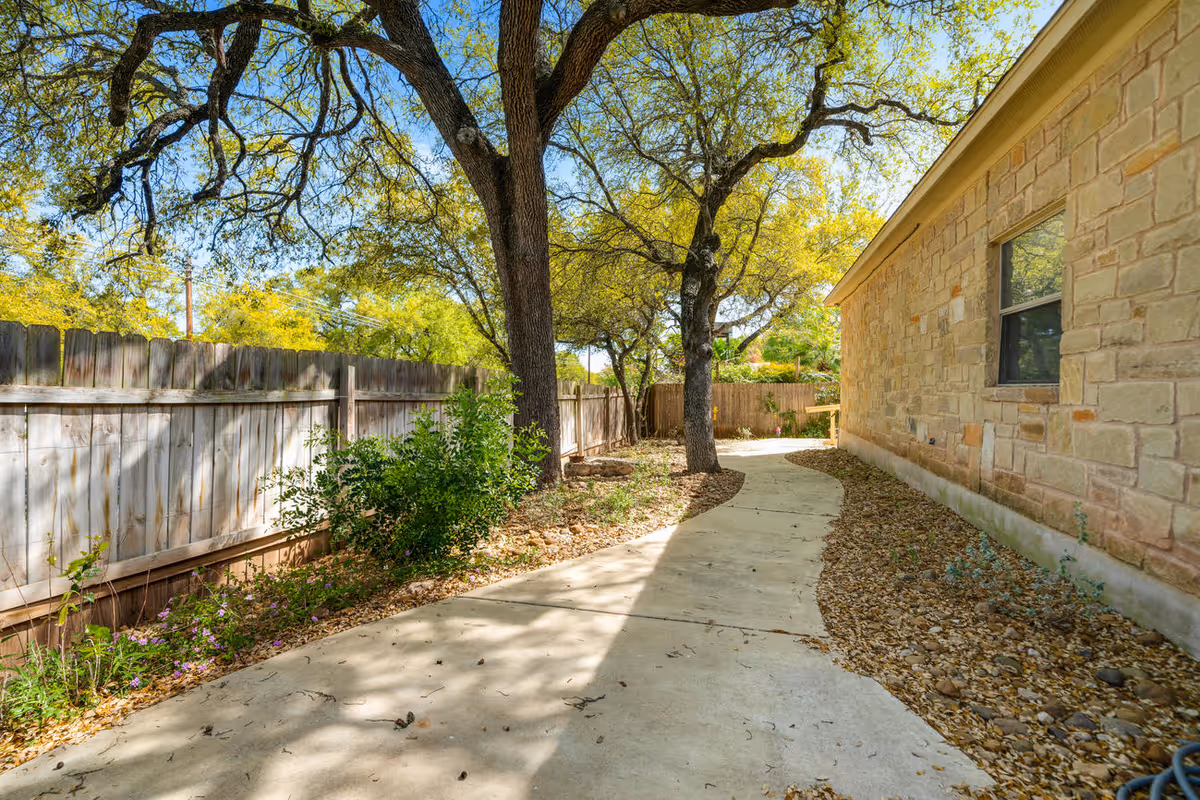 A winding concrete pathway runs alongside a stone building with a window. The path is bordered by a wooden fence on one side and landscaped areas with trees, bushes, and rocks on both sides. Sunlight filters through the tree branches, casting shadows on the path.