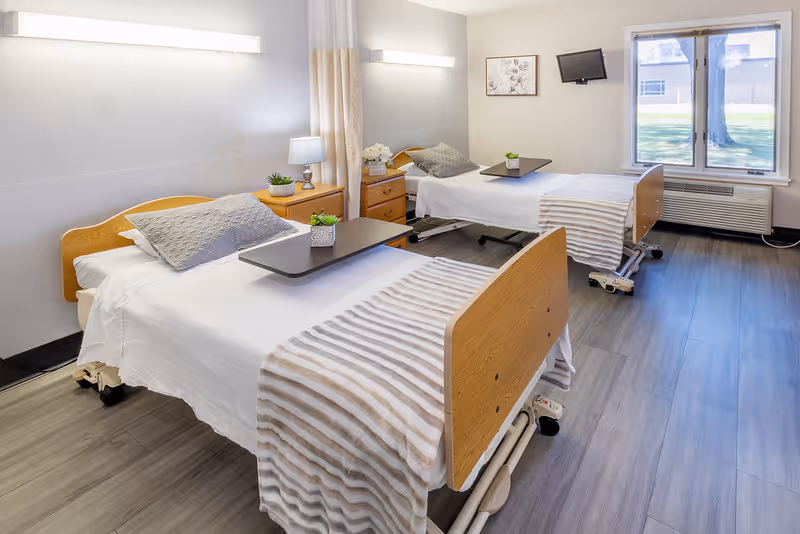 A bright and clean room with two hospital-style beds, each with a gray pillow and a striped blanket. Each bed has an overbed table with a small potted plant. Between the beds is a wooden nightstand with a lamp and decorative items. A window with a view of the outdoors lets in natural light, and a small wall-mounted TV is visible. The floor is covered with wood-like vinyl flooring.