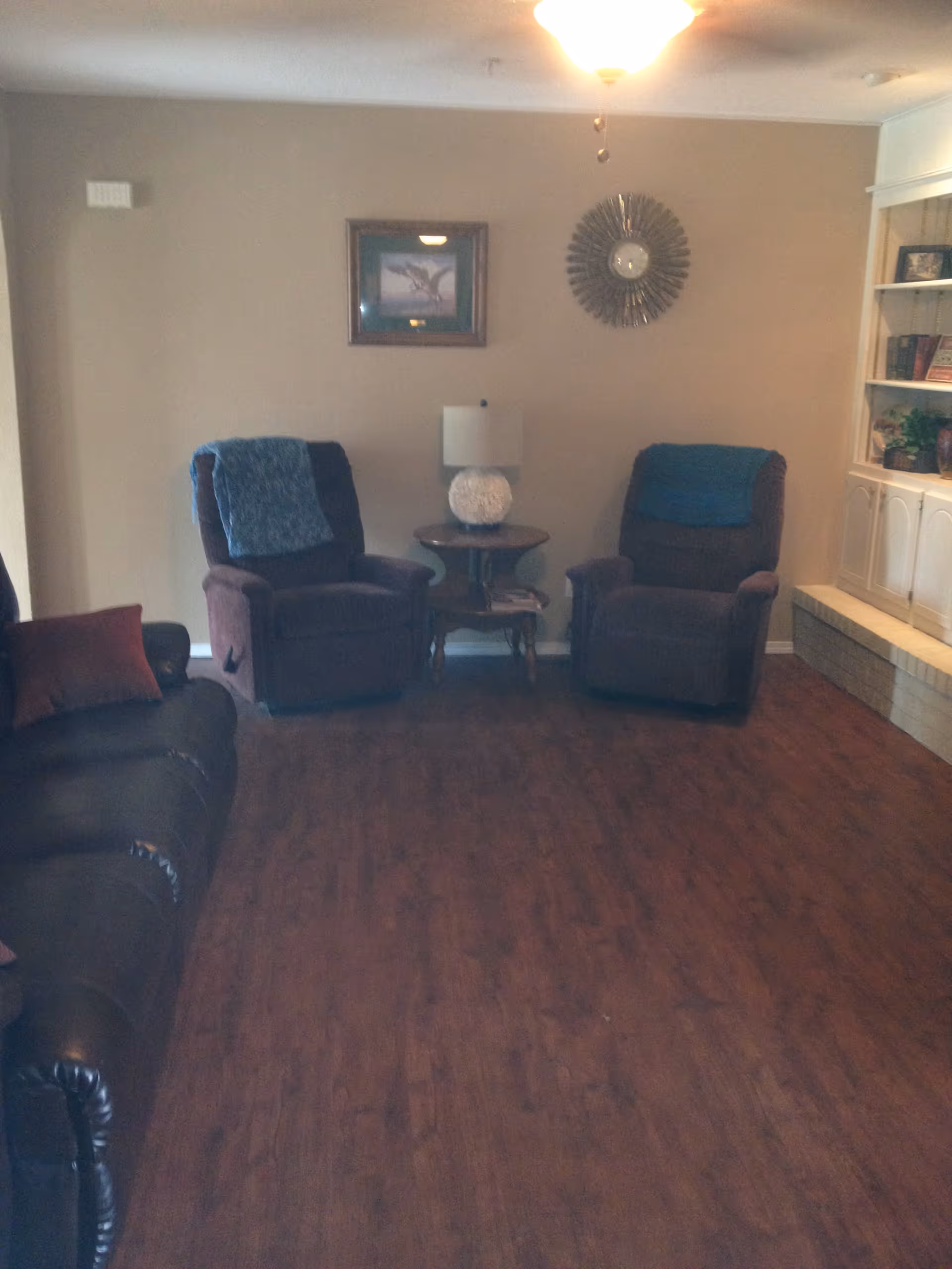 A living room with two brown recliner chairs with blue throws, a brown leather sofa with a red pillow, a round wooden side table with a white lamp, a wall clock, a framed picture, and built-in white shelves with books and plants.