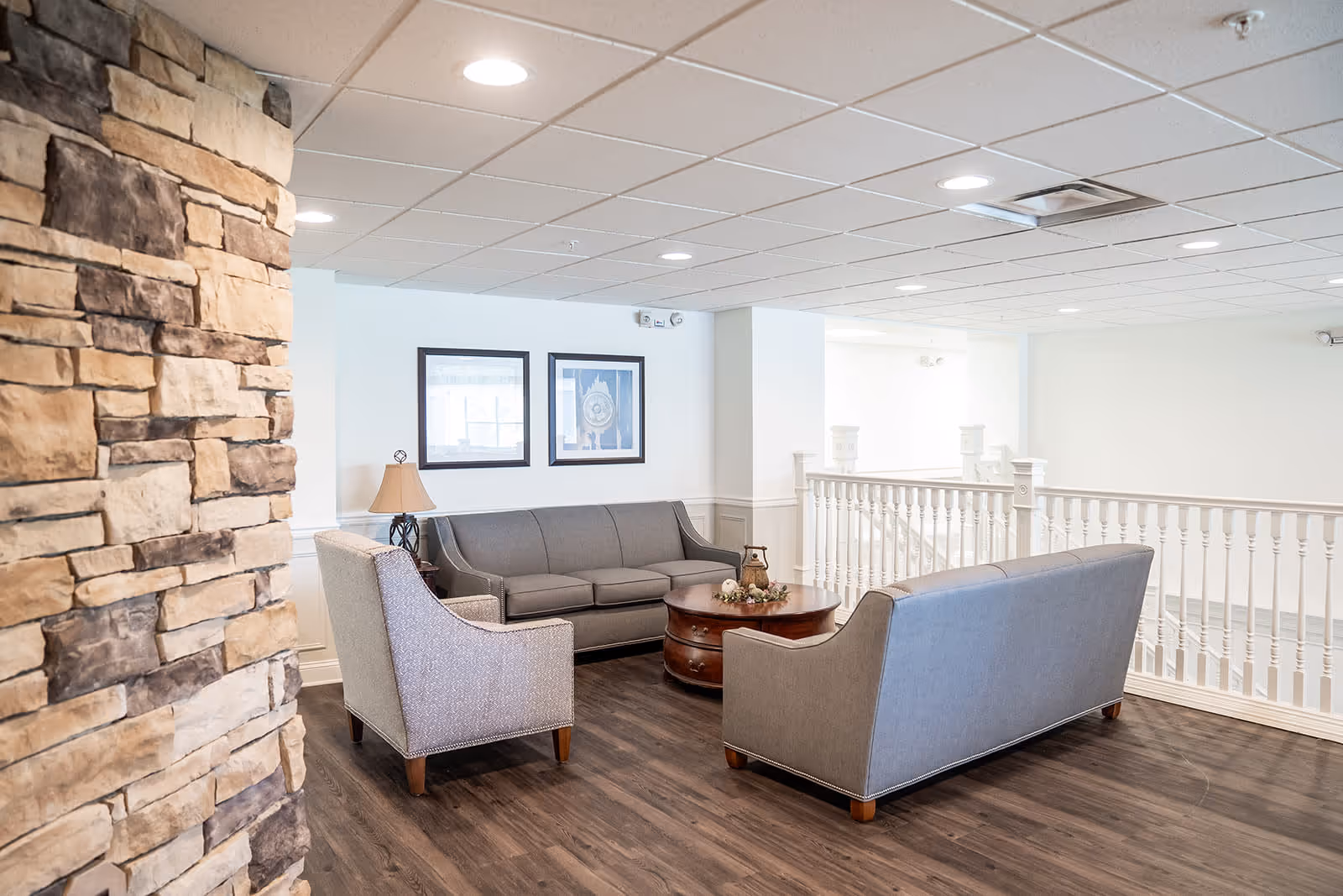 A bright and spacious sitting area with two gray sofas and a gray armchair arranged around a round wooden coffee table. The room features a stone accent wall on the left, white walls with two framed pictures, a table lamp, and a white railing overlooking a lower level. The floor is dark wood, and the ceiling has recessed lighting.