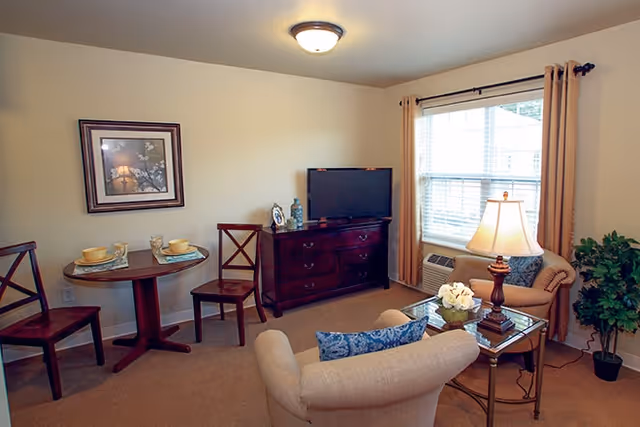 A cozy living room with beige walls and carpet. The room features a small round dining table with two chairs, a wooden dresser with a flat-screen TV on top, and two upholstered armchairs with a glass coffee table between them. A table lamp and a potted plant are also visible near a window with beige curtains.