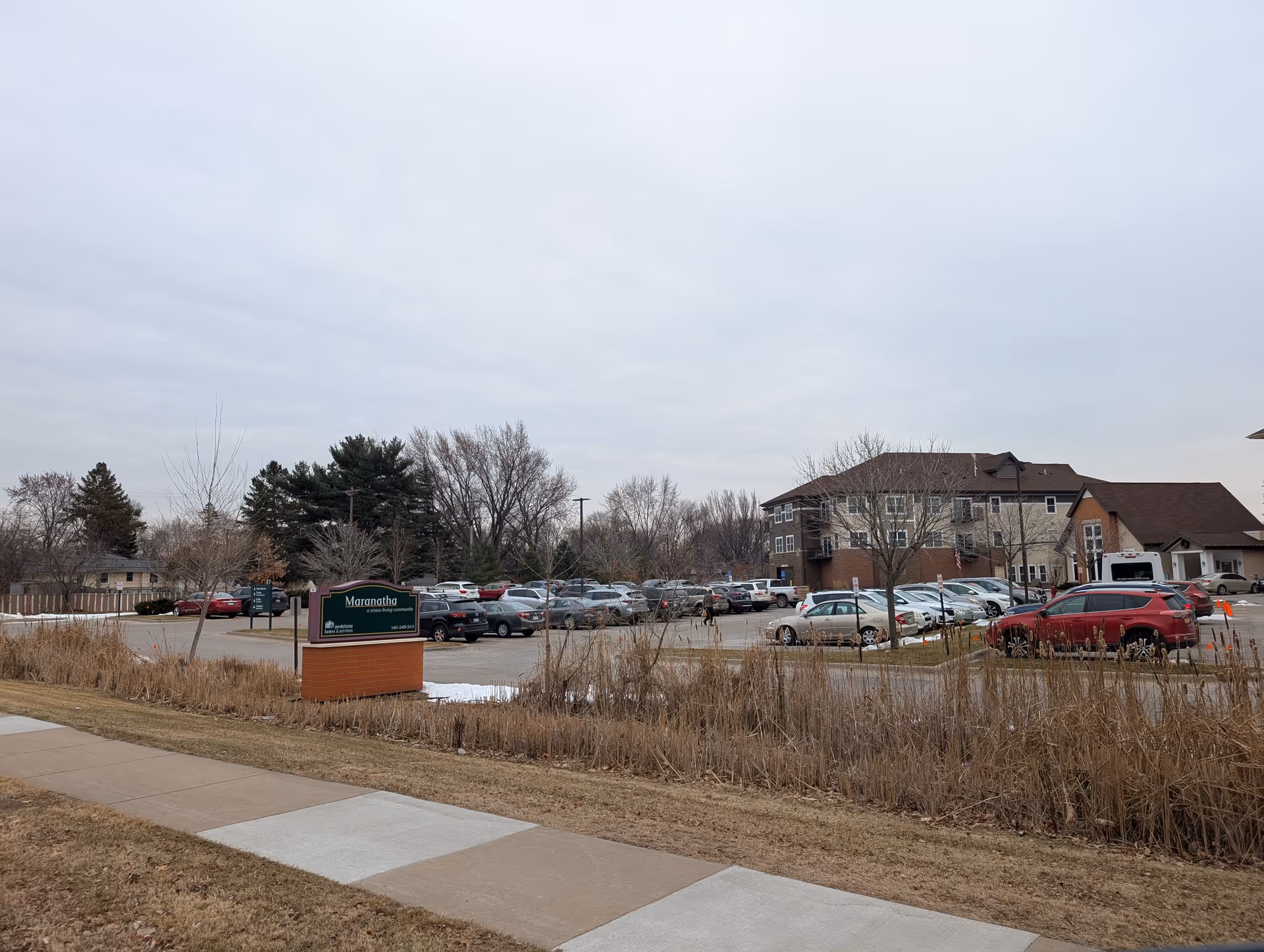 Exterior view of Maranatha Place senior living facility showing a parking lot with multiple cars, a sidewalk, dry grass, leafless trees, and a cloudy sky.