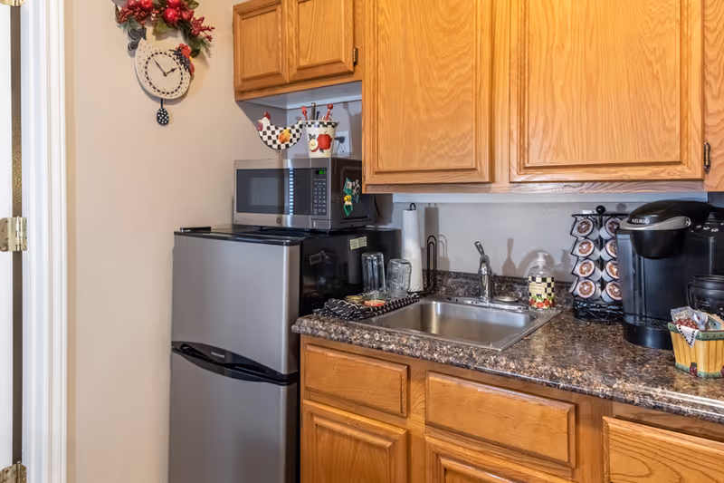 Small kitchen area with a mini refrigerator, microwave, sink, Keurig coffee maker, and wooden cabinets.