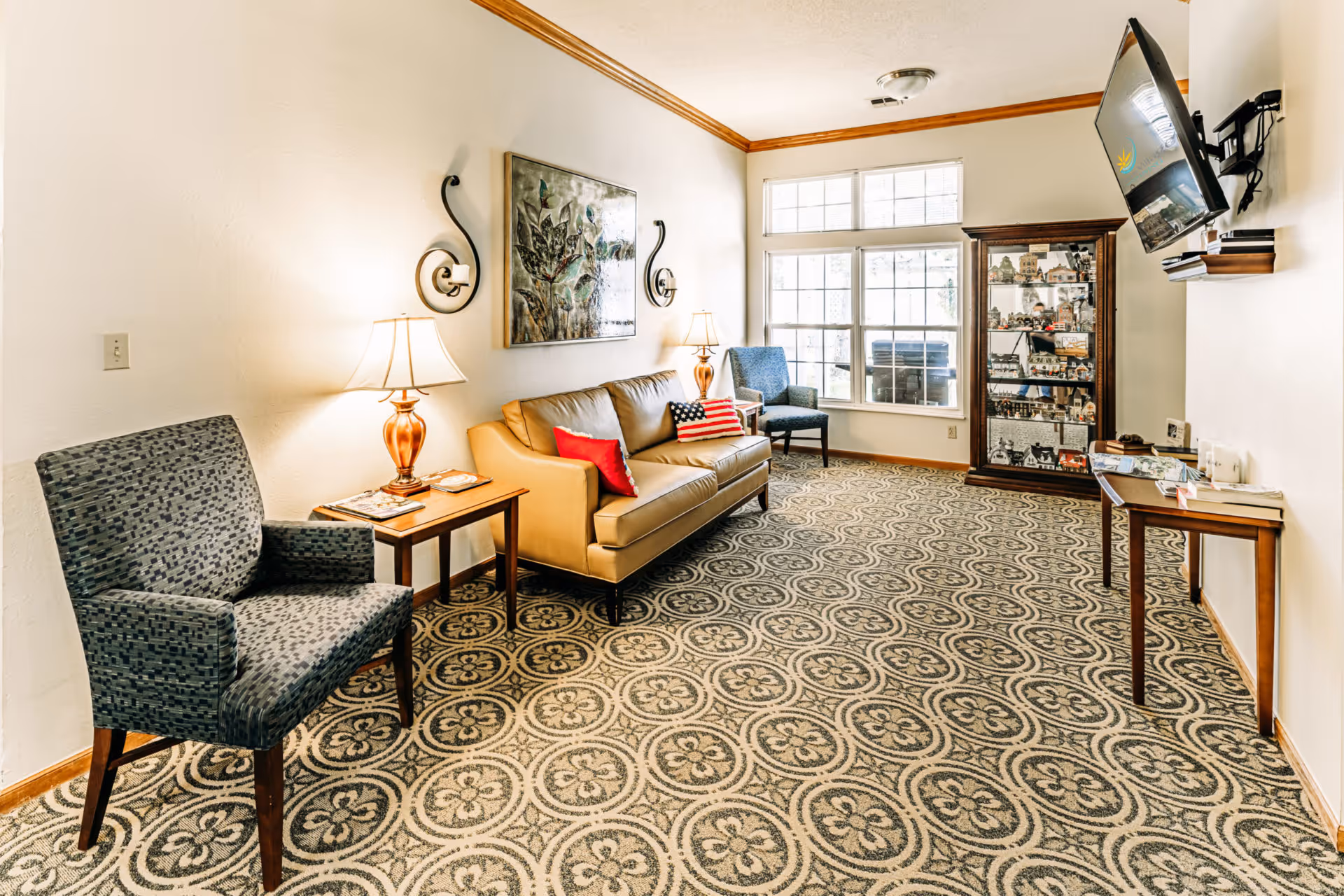 A well-lit living room with a patterned carpet, a tan leather sofa with red and American flag pillows, two blue upholstered chairs, a wooden side table with a lamp, a wall-mounted TV, a glass display cabinet filled with miniature houses, and large windows letting in natural light.