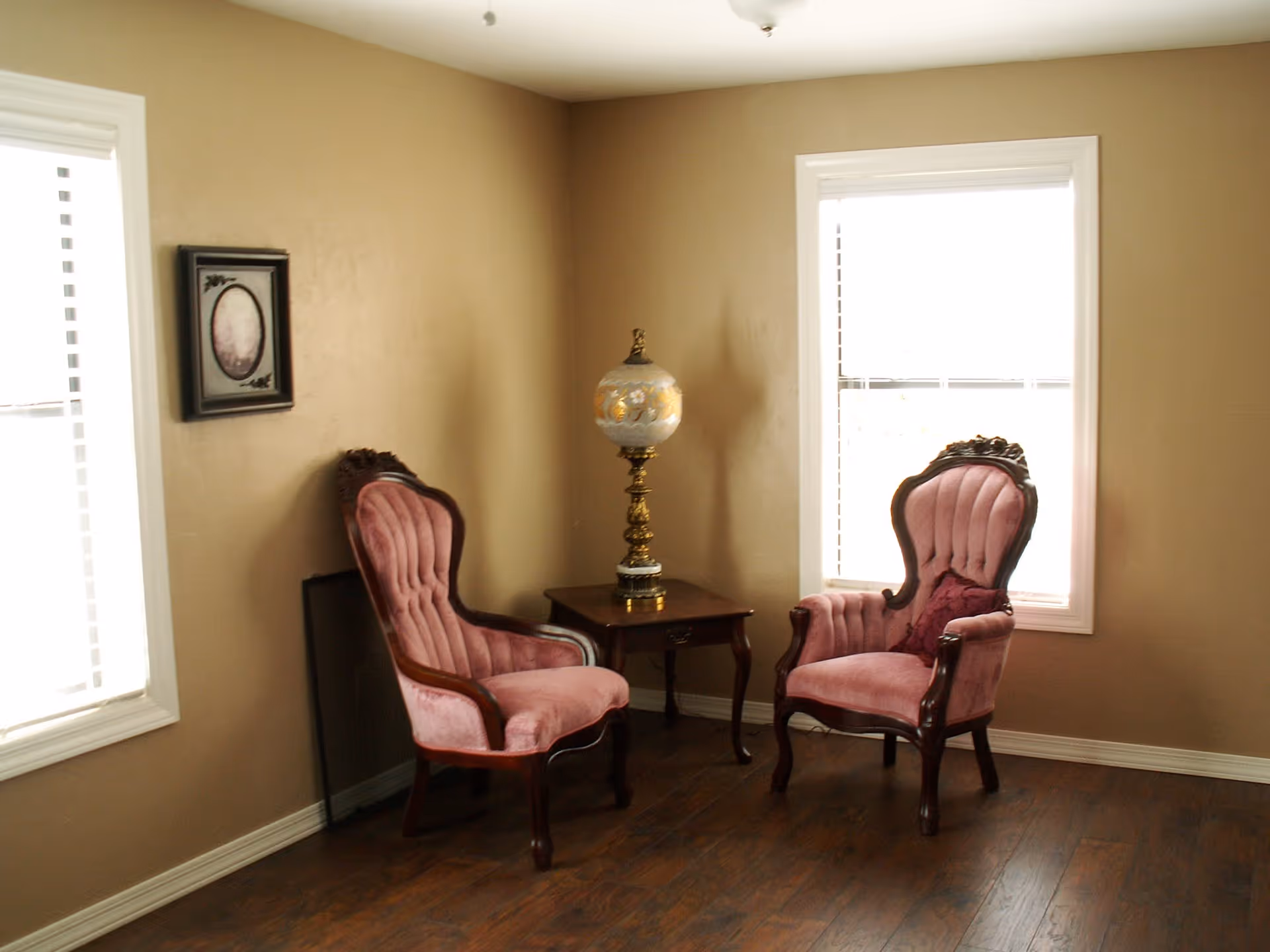 A cozy corner of a room with two vintage-style pink upholstered armchairs facing each other, separated by a small wooden side table with an ornate lamp on it. The room has beige walls, two windows with white blinds, and dark wooden flooring. A framed picture hangs on the wall beside one of the windows.