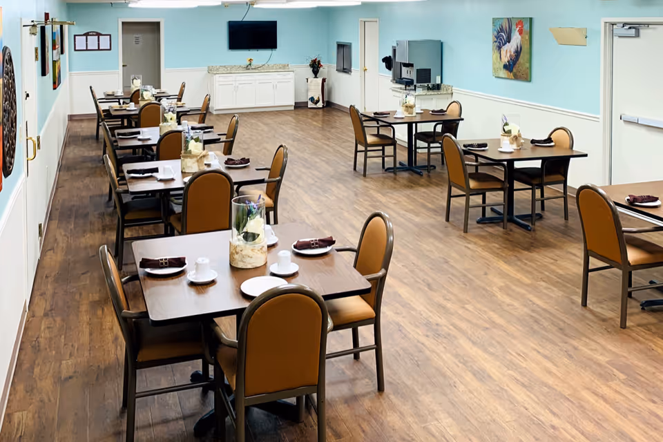 Communal dining room with multiple set tables and chairs, a TV on the far wall and a beverage station against a light blue wall.