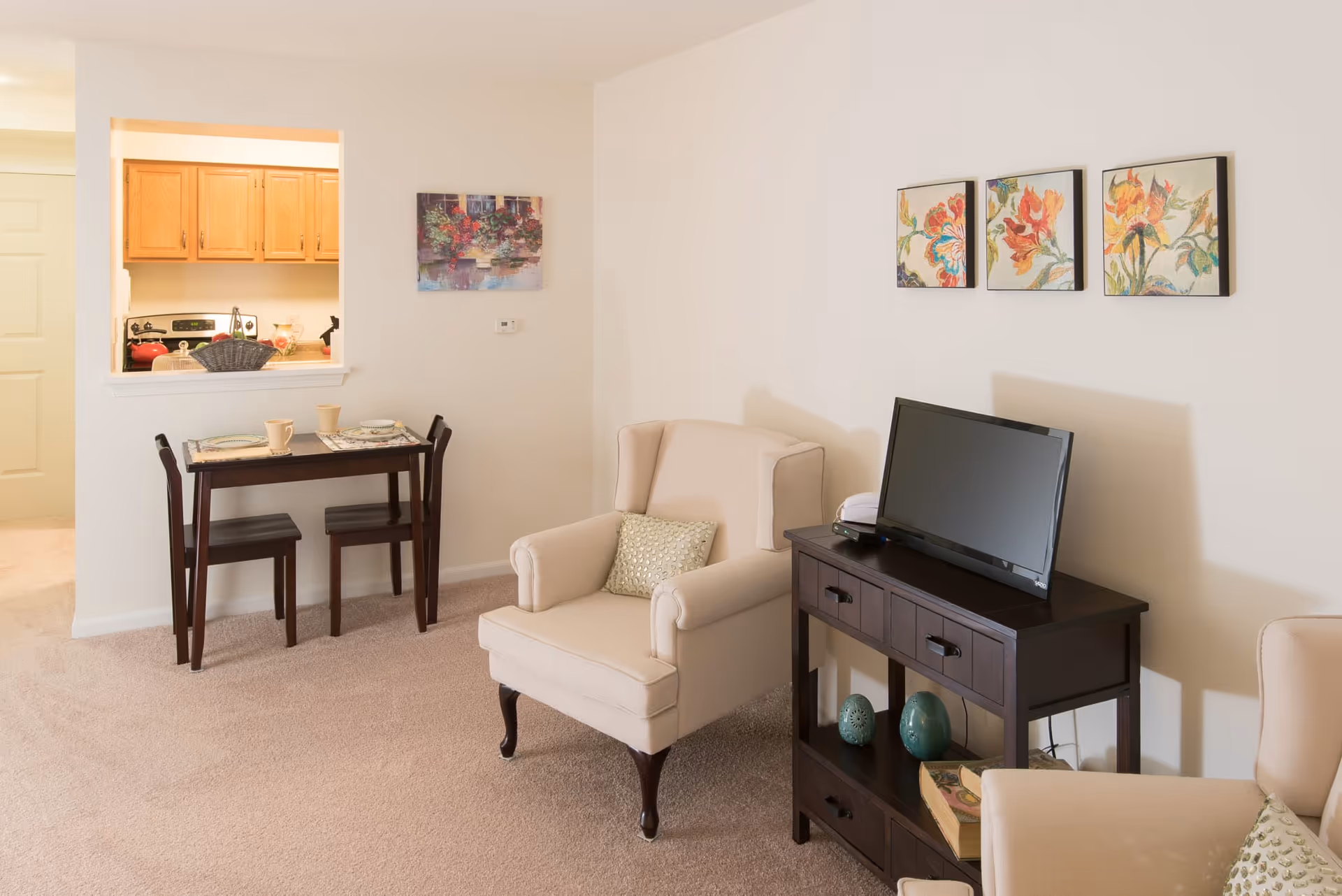 Cozy living room with two upholstered armchairs, a TV on a dark wood console, a small dining table and a pass-through to the kitchen.