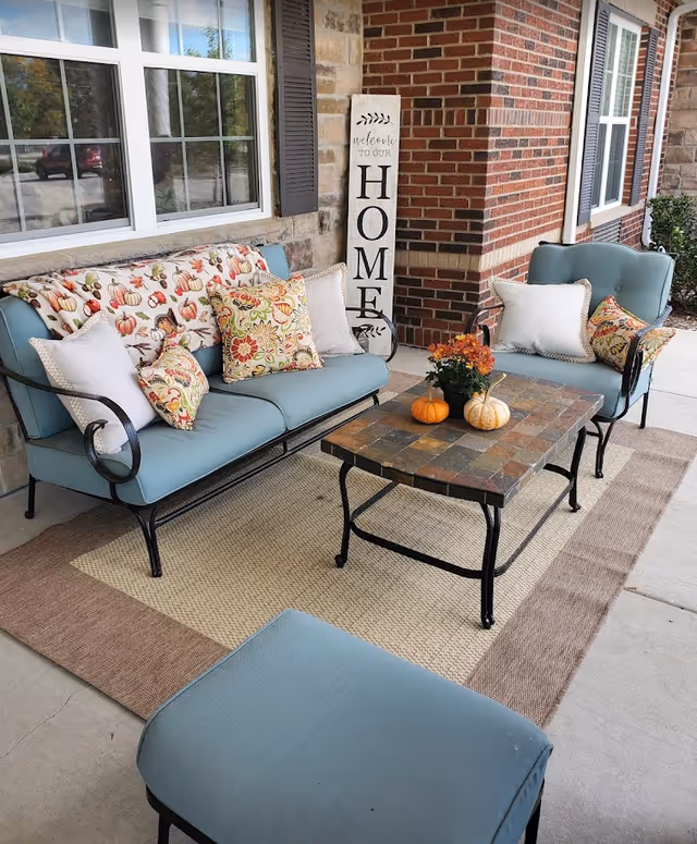 Outdoor seating area with light blue cushioned metal furniture including a loveseat, chair, and ottoman on a beige and brown rug. The loveseat has decorative pillows with floral and pumpkin patterns. A small square table with a tiled top holds two small pumpkins and a flower arrangement. A vertical wooden sign next to the brick wall reads 'welcome to our HOME'.