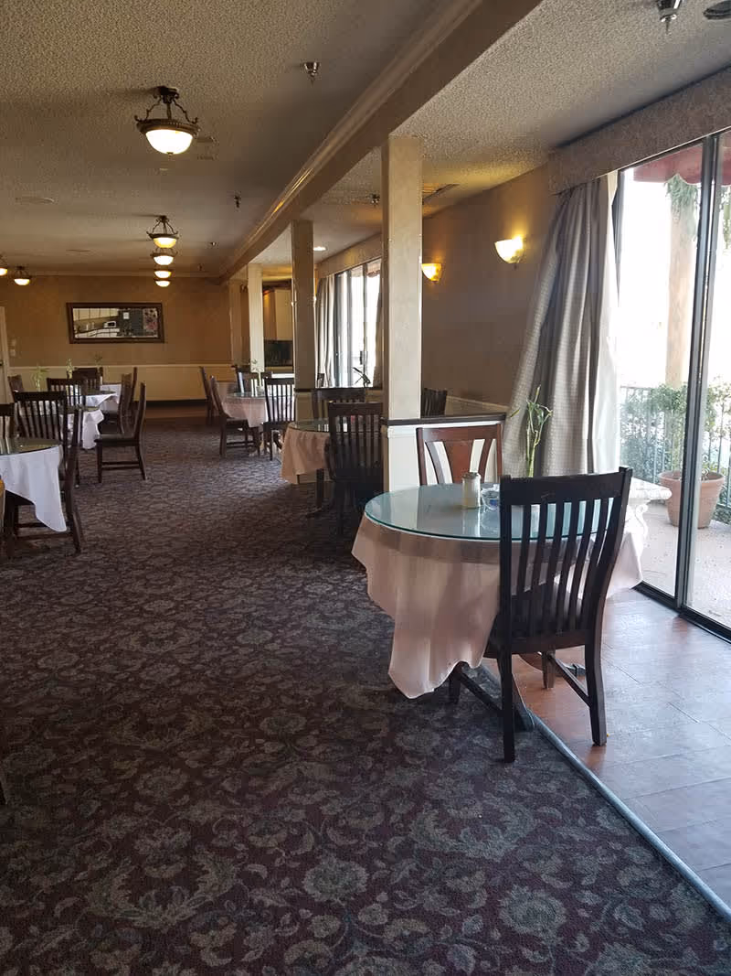 Interior view of a dining room with multiple round tables covered with white tablecloths and glass tops, each surrounded by wooden chairs. The room has patterned carpet flooring, beige walls, ceiling lights, and large windows with curtains letting in natural light. There are some potted plants visible outside the windows.
