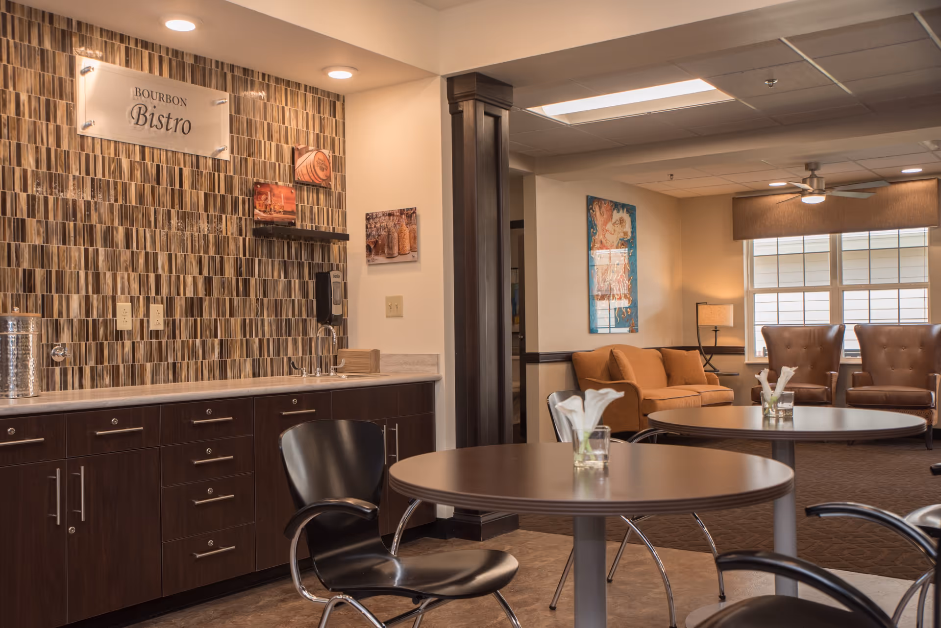 Cozy communal bistro area with round tables and chairs, a serving counter with a tiled backsplash and 'Bourbon Bistro' sign, and a seating area with a sofa and armchairs.