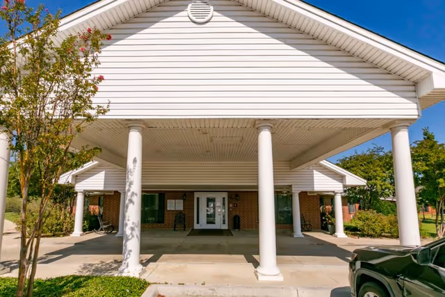 Front exterior view of a single-story building with white siding and a covered entrance supported by four white columns. There are trees and shrubs on either side of the entrance, and a black vehicle is partially visible on the right side of the image under a clear blue sky.