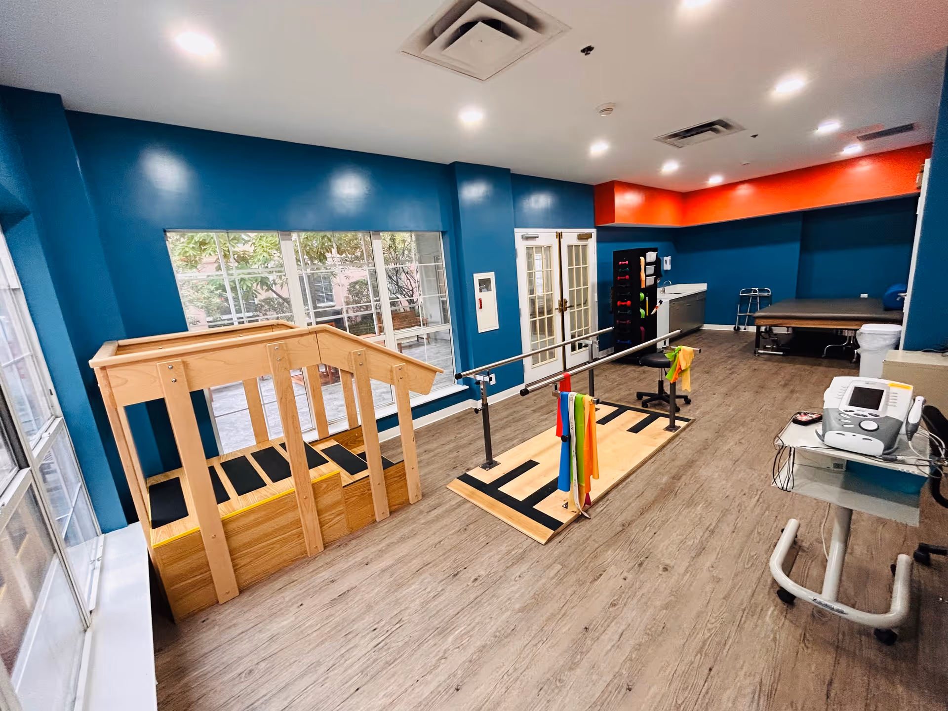 A physical therapy room with wooden stairs for rehabilitation, parallel bars with colorful resistance bands, exercise equipment, and a treatment table. The walls are painted blue with an orange accent near the ceiling, and large windows allow natural light to enter the room.