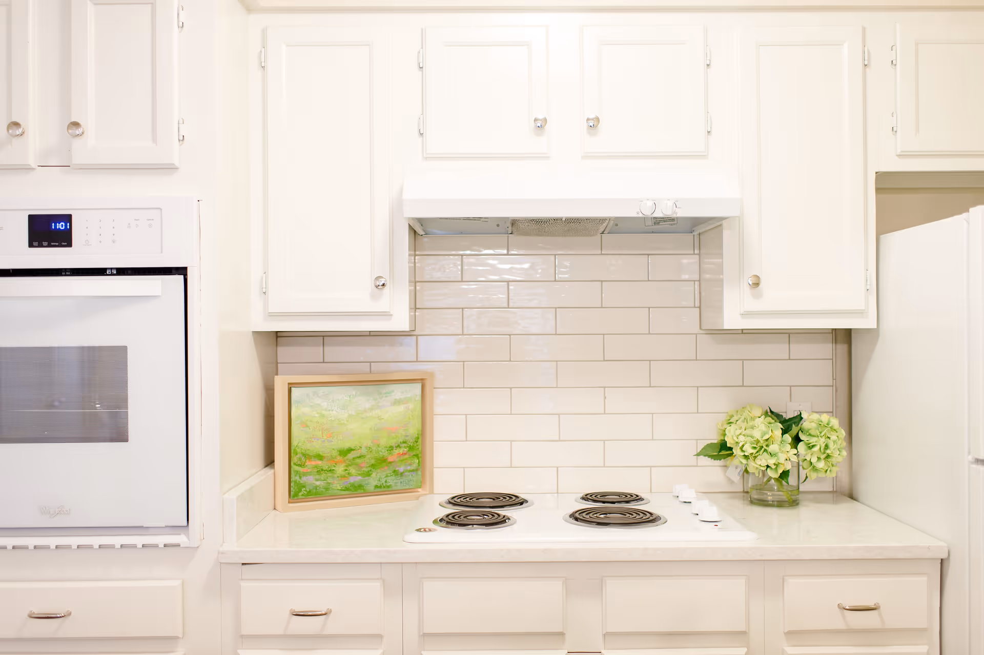 Bright white kitchen with a stovetop and oven, subway tile backsplash, upper cabinets, a small framed painting and a vase of green flowers on the counter.