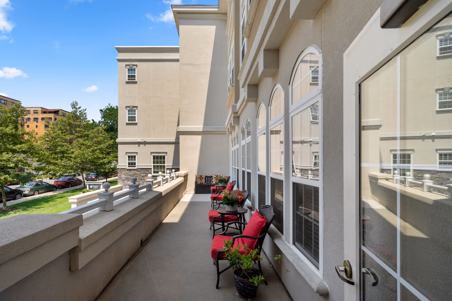 Outdoor balcony area at Symphony Square with several black metal chairs featuring red cushions, small tables with potted plants, and a cushioned bench at the far end. The balcony overlooks a parking lot and green trees under a clear blue sky.