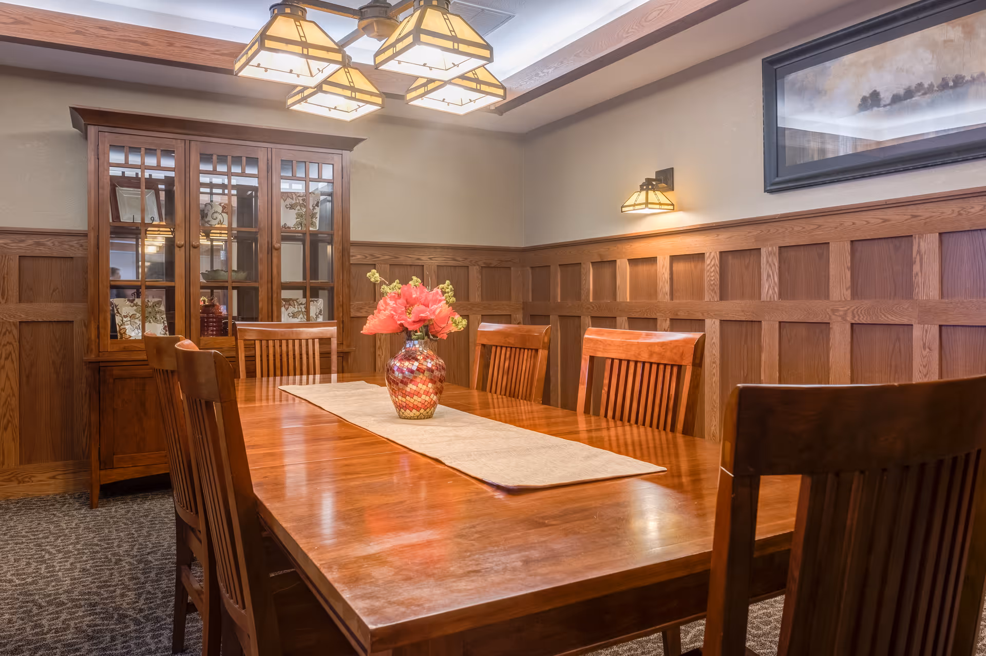 A warm, wood-paneled dining room with a long wooden table surrounded by wooden chairs. A decorative vase with pink flowers sits on a beige table runner in the center of the table. A wooden cabinet with glass doors is against the wall, and a framed artwork hangs above the wood paneling. Soft lighting comes from a ceiling fixture and a wall sconce.