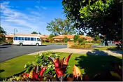 View of the exterior grounds of The Meadows At Cypress Gardens facility, showing a driveway with a white shuttle bus parked near a single-story building with a red-tiled roof, surrounded by green lawns, trees, and colorful plants in the foreground under a clear blue sky.