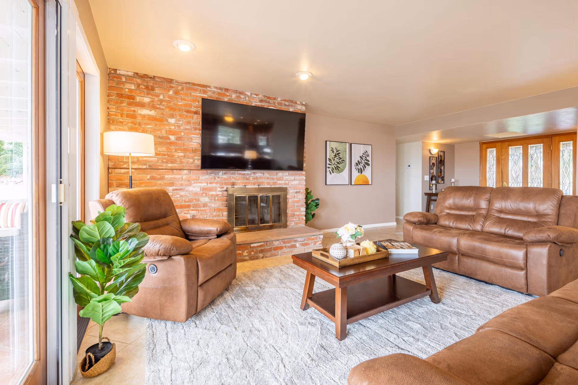 A cozy living room with brown leather recliner chairs and a sofa arranged around a wooden coffee table on a light gray rug. A brick fireplace with a mounted flat-screen TV is on the wall, and there are two framed botanical prints on the adjacent wall. A floor lamp and a potted plant are near the sliding glass door that leads outside.