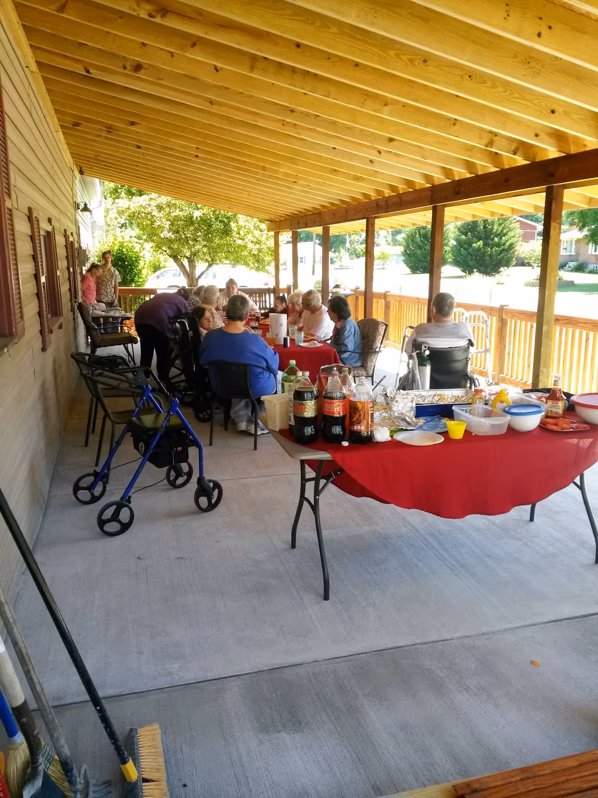 A group of seniors seated around tables under a covered wooden porch sharing a meal, with food and drinks on a red-covered table in the foreground.