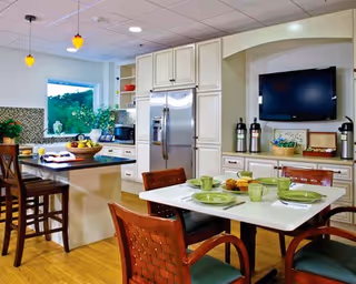 A bright and modern kitchen and dining area in a senior living facility. The kitchen features white cabinetry, a stainless steel refrigerator, a black countertop island with two wooden bar stools, and a window with a view of greenery outside. The dining area has a white table set with green plates, cups, and utensils, surrounded by four wooden chairs with green cushions. A flat-screen TV is mounted on the wall above a countertop with decorative items and coffee dispensers.